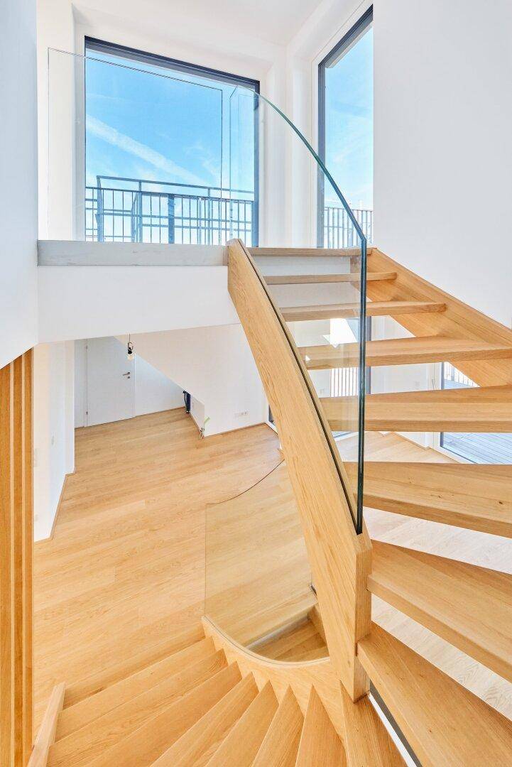 Elegante Holztreppe mit Glasgeländer in einem hellen Treppenhaus, das viel Tageslicht hereinlässt.