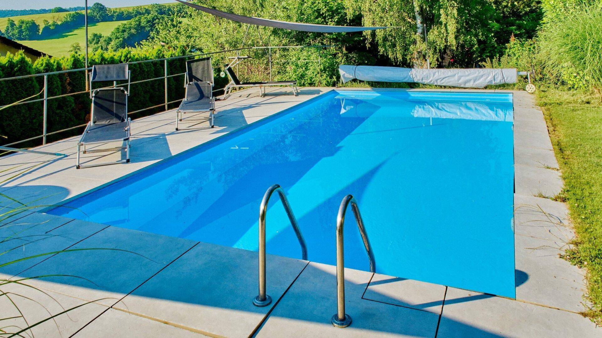 Rechteckiger Außenpool mit blauer Auskleidung, umgeben von einer gepflasterten Terrasse und Liegestühlen.