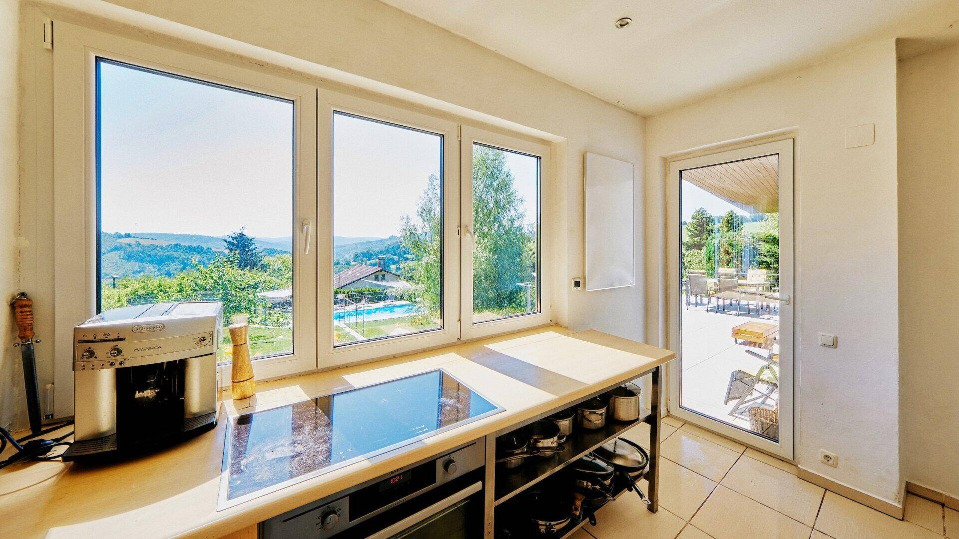 Helle Küche mit Kochfeld, Kaffeemaschine und Panoramafenstern mit Blick auf den Pool und die Landschaft.
