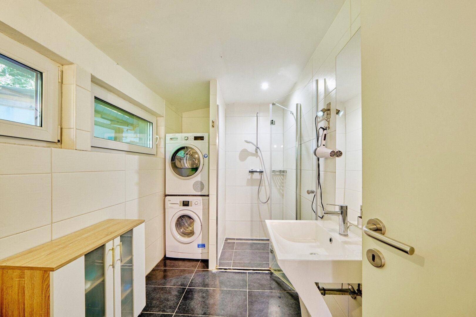 Modernes Badezimmer mit Dusche, Waschmaschine und Trockner, sowie einem Fenster für Tageslicht.