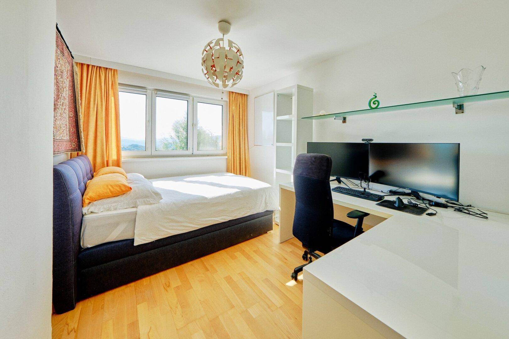Schlafzimmer mit Holzboden, Schreibtisch mit zwei Monitoren und Blick ins Grüne.