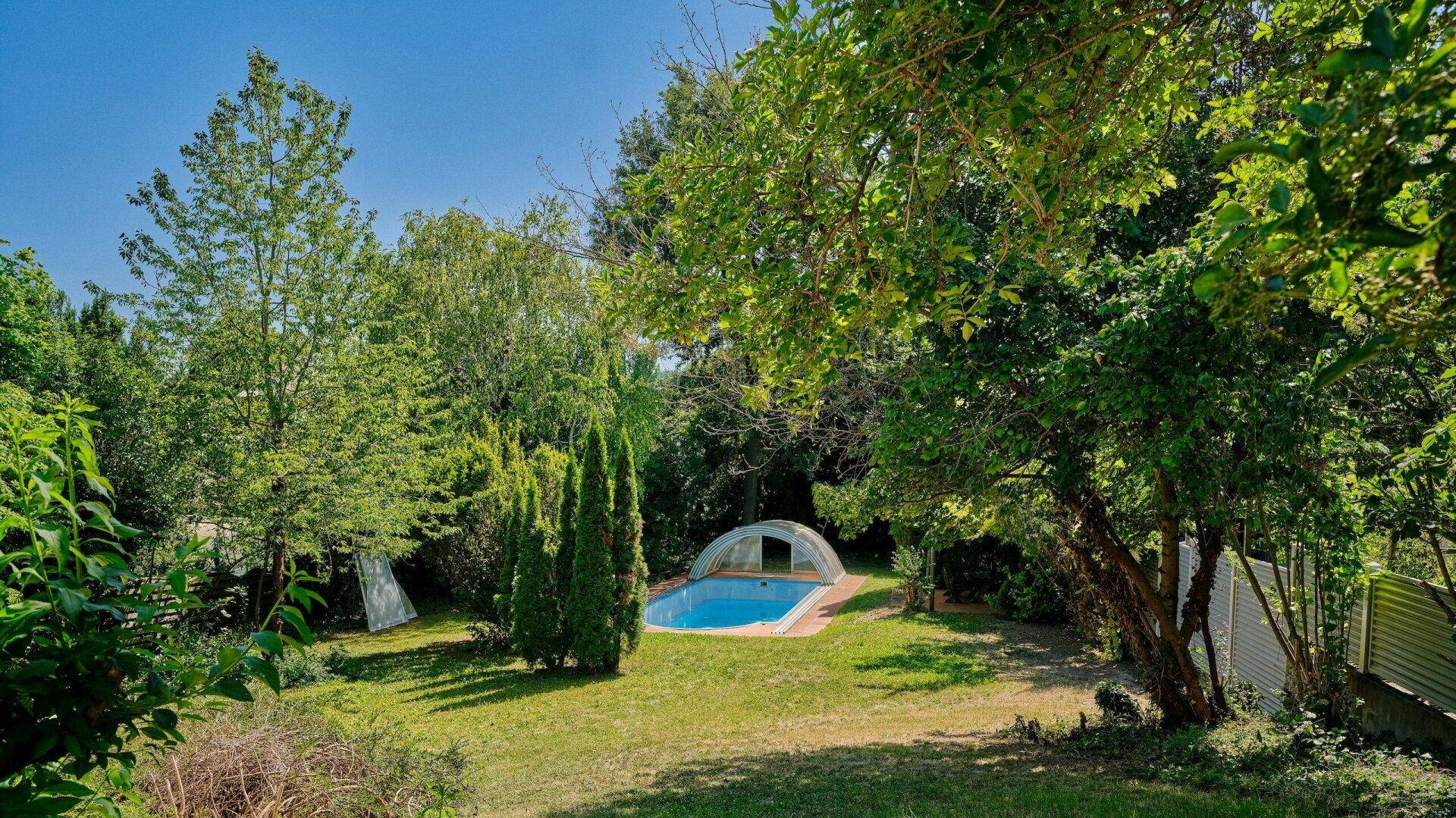 Weitläufiger Garten mit Rasenfläche, altem Baumbestand und einem überdachten Poolbereich.