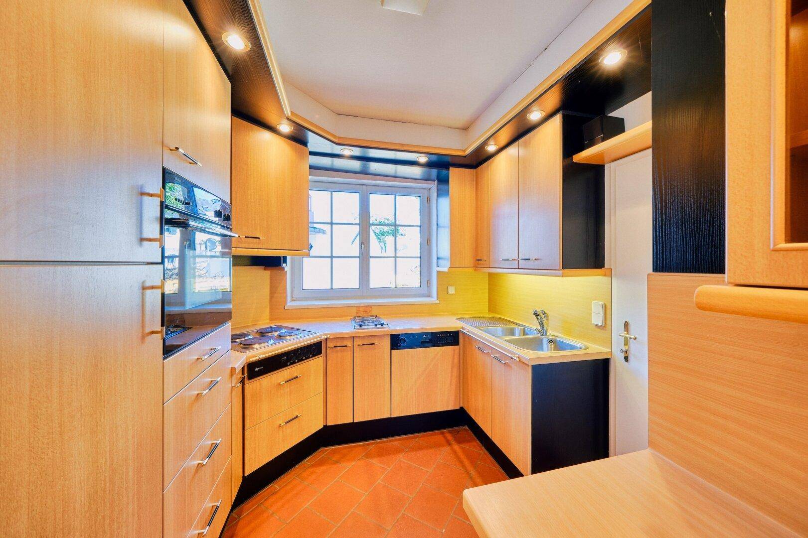 Funktionale Küche mit Holzfronten, Einbaugeräten und Terrakotta-Fliesenboden, gut beleuchtet.