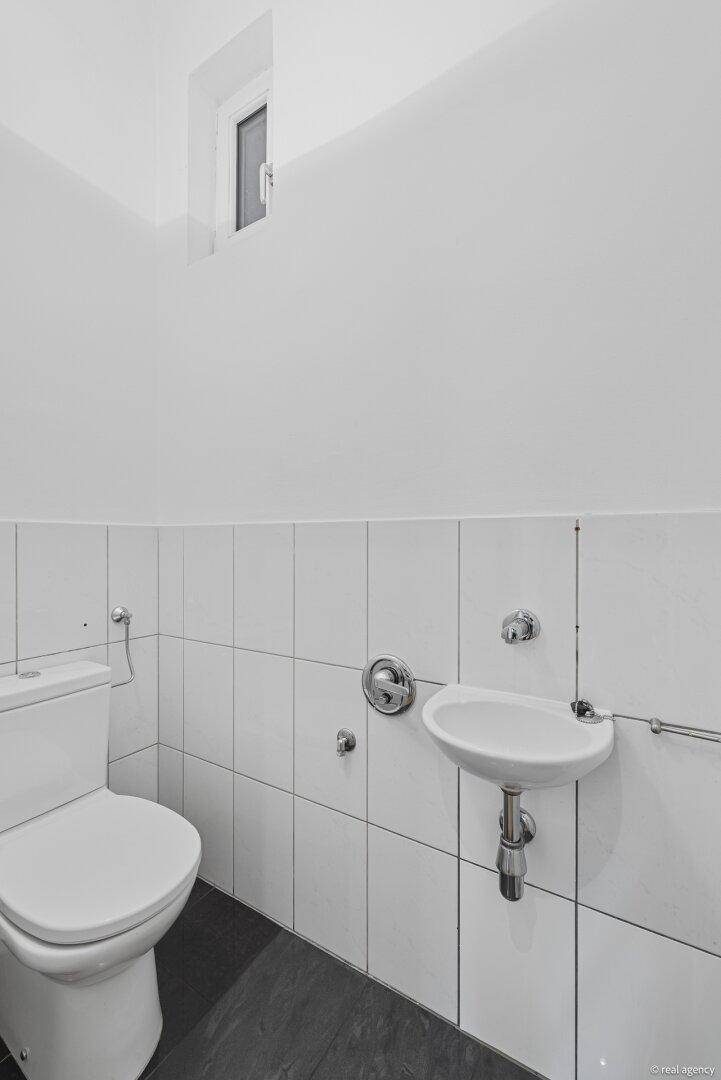 Kleine Gästetoilette mit weiß gefliesten Wänden, dunklem Boden und einem Fenster.