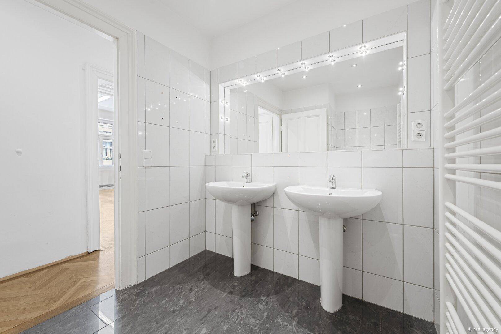 Helles Badezimmer mit zwei Waschbecken, großem Spiegel und weiß gefliesten Wänden.