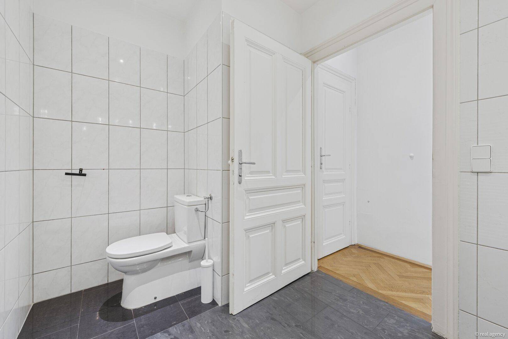 Badezimmer mit Toilette, weiß gefliesten Wänden und dunklem Boden, angrenzend an einen anderen Raum.
