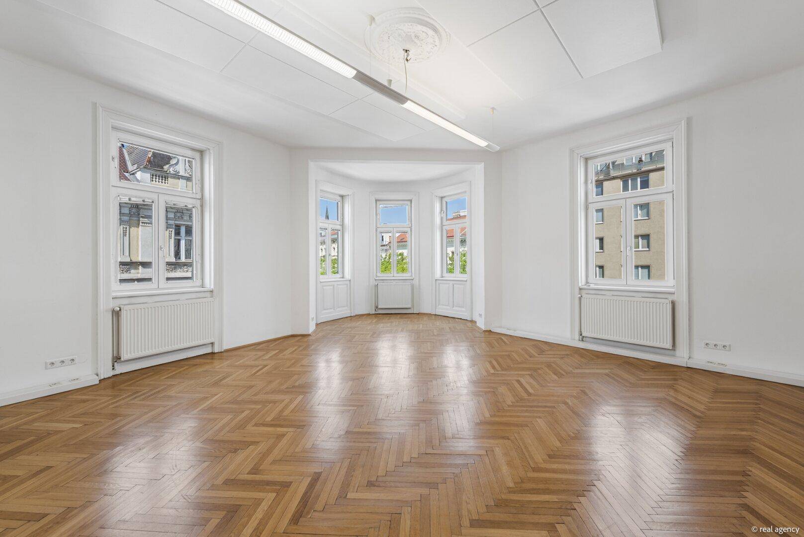 Geräumiger Raum mit Fischgrätparkett, hohen Decken und großen Fenstern, bietet viel Tageslicht.