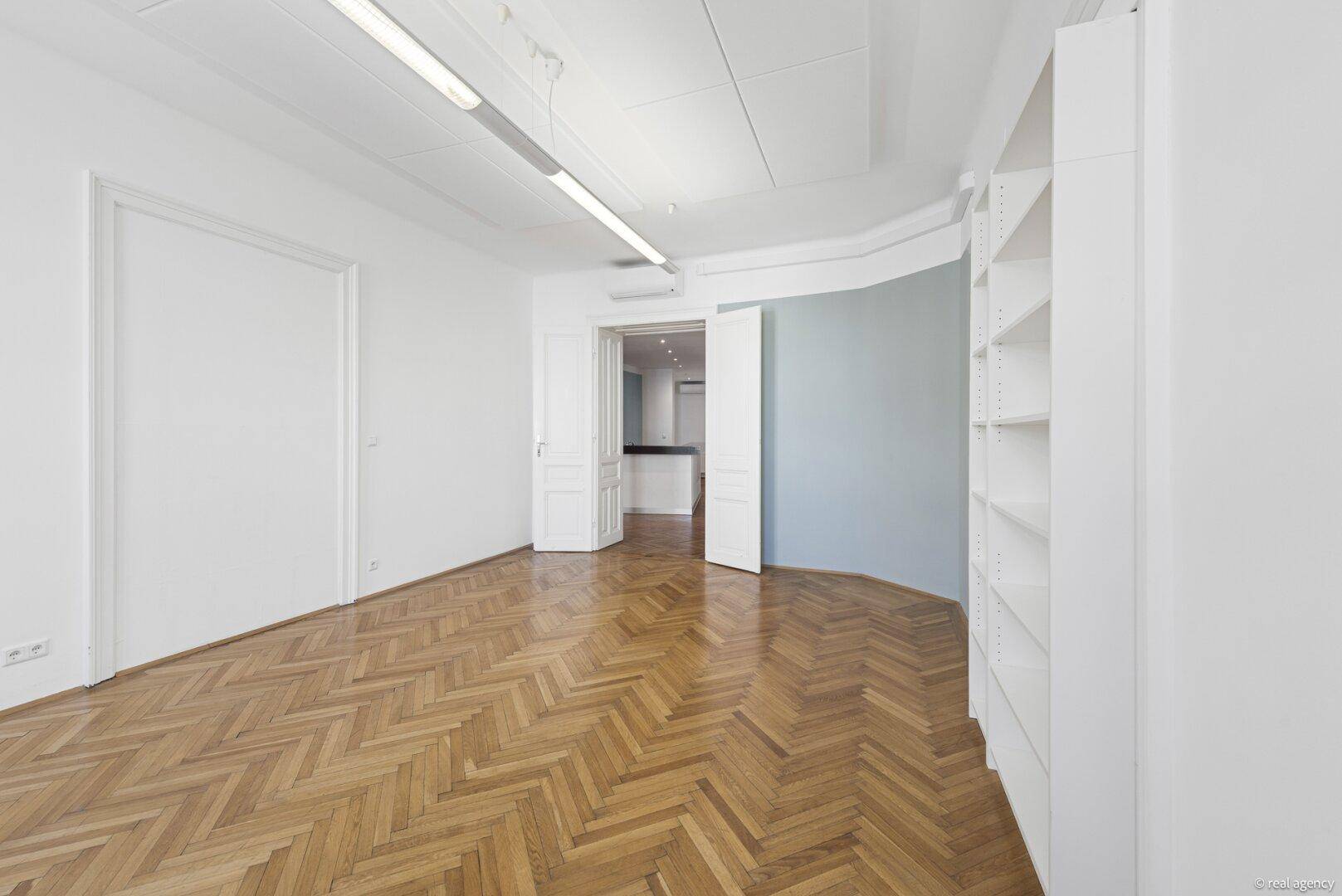 Heller Raum mit Fischgrätparkett, weiß gestrichenen Wänden und Blick in angrenzende Zimmer.