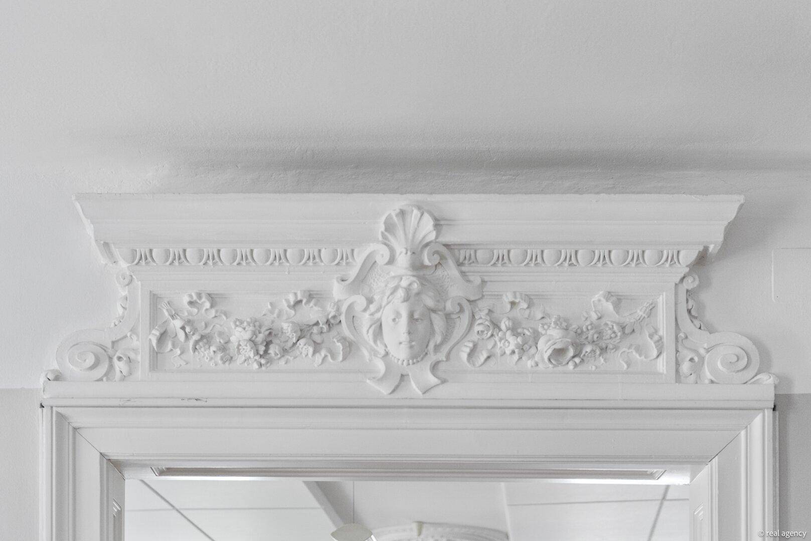 Symmetrisches Stuckrelief mit zentralem Medaillon über einem klassischen weißen Türrahmen.