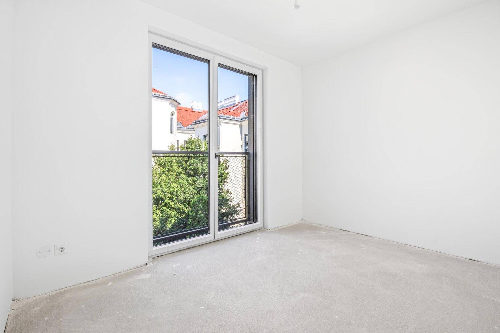 Unmöblierter Raum mit weiß gestrichenen Wänden und großem Fenster, das einen Blick auf Bäume und Dächer bietet.