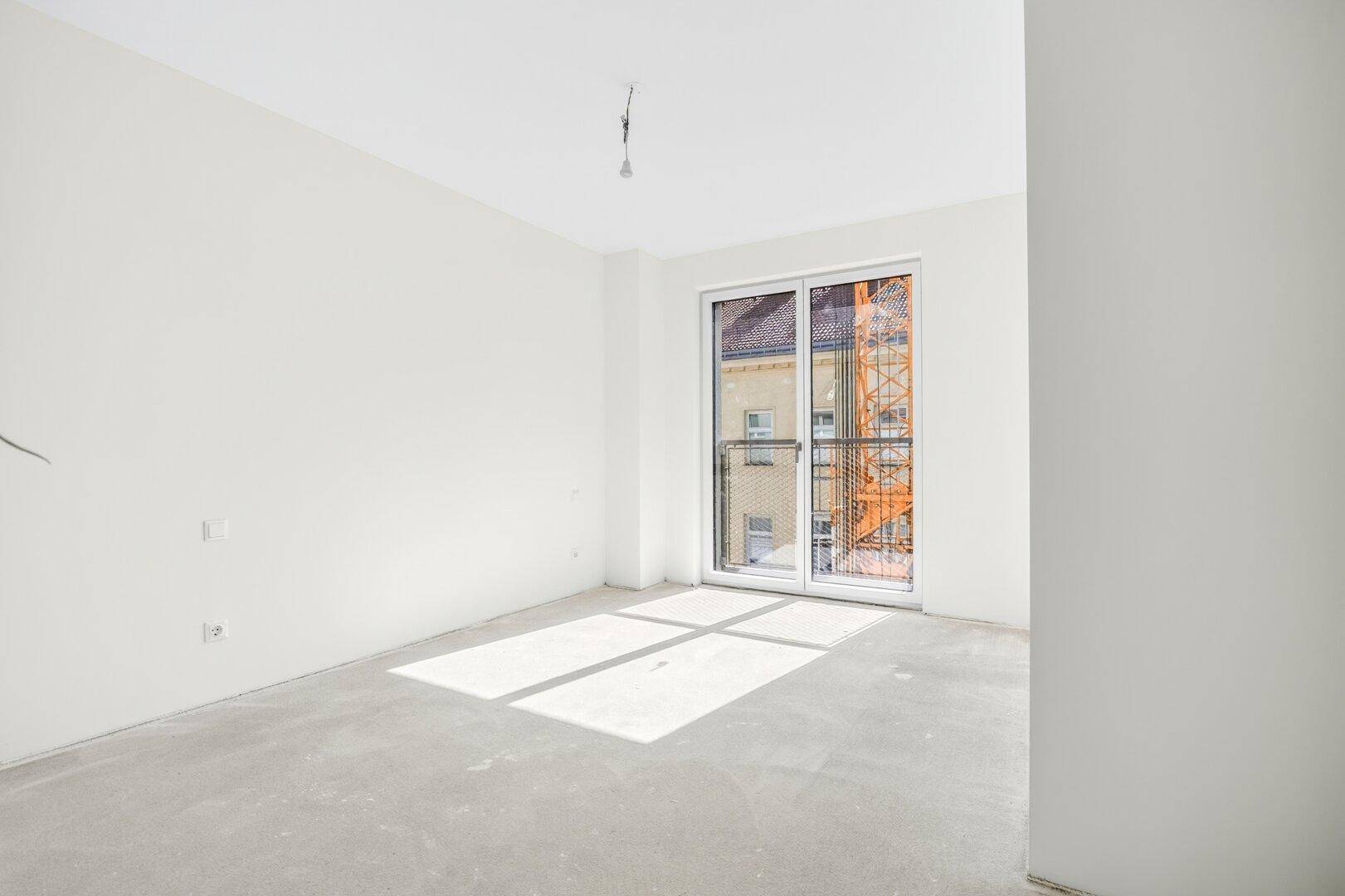 Heller Raum mit Sonnenlicht und großem Fenster mit französischem Balkon, Blick auf ein Gebäude und einen Baukran.