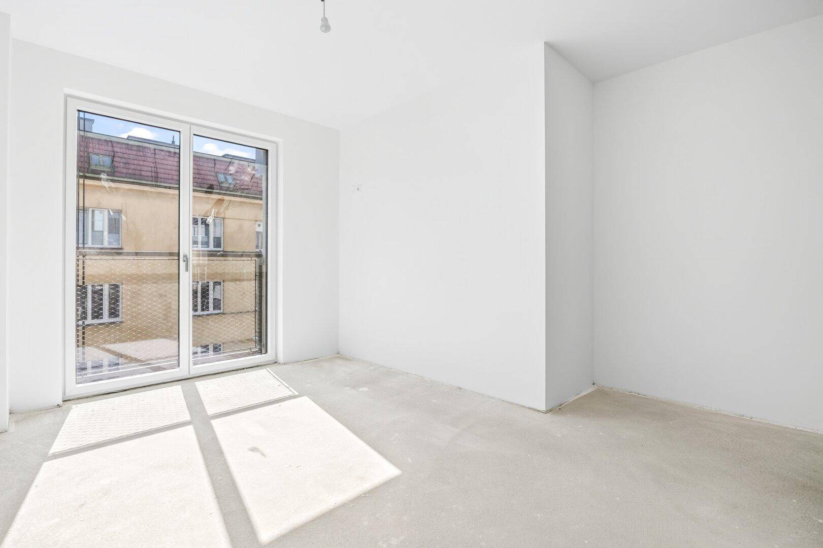 Heller Raum mit großem Fenster und französischem Balkon, der viel Sonnenlicht auf den Rohbetonboden wirft.