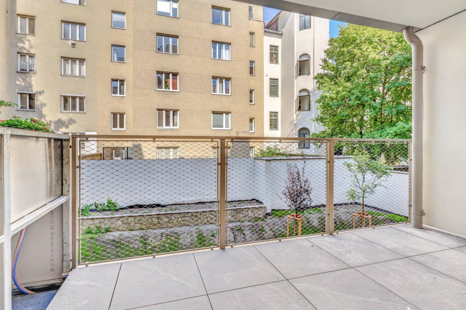 Geräumiger Balkon mit Fliesen und modernem Geländer, Blick auf die Fassade eines Mehrfamilienhauses und einen kleinen Garten.