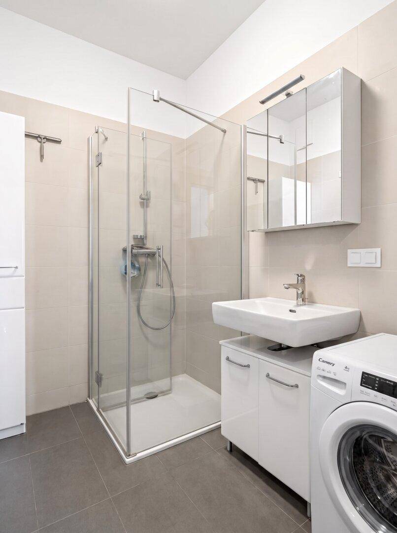 Modernes Badezimmer mit ebenerdiger Dusche, Einzelwaschbecken und Waschmaschine, hell gefliest.