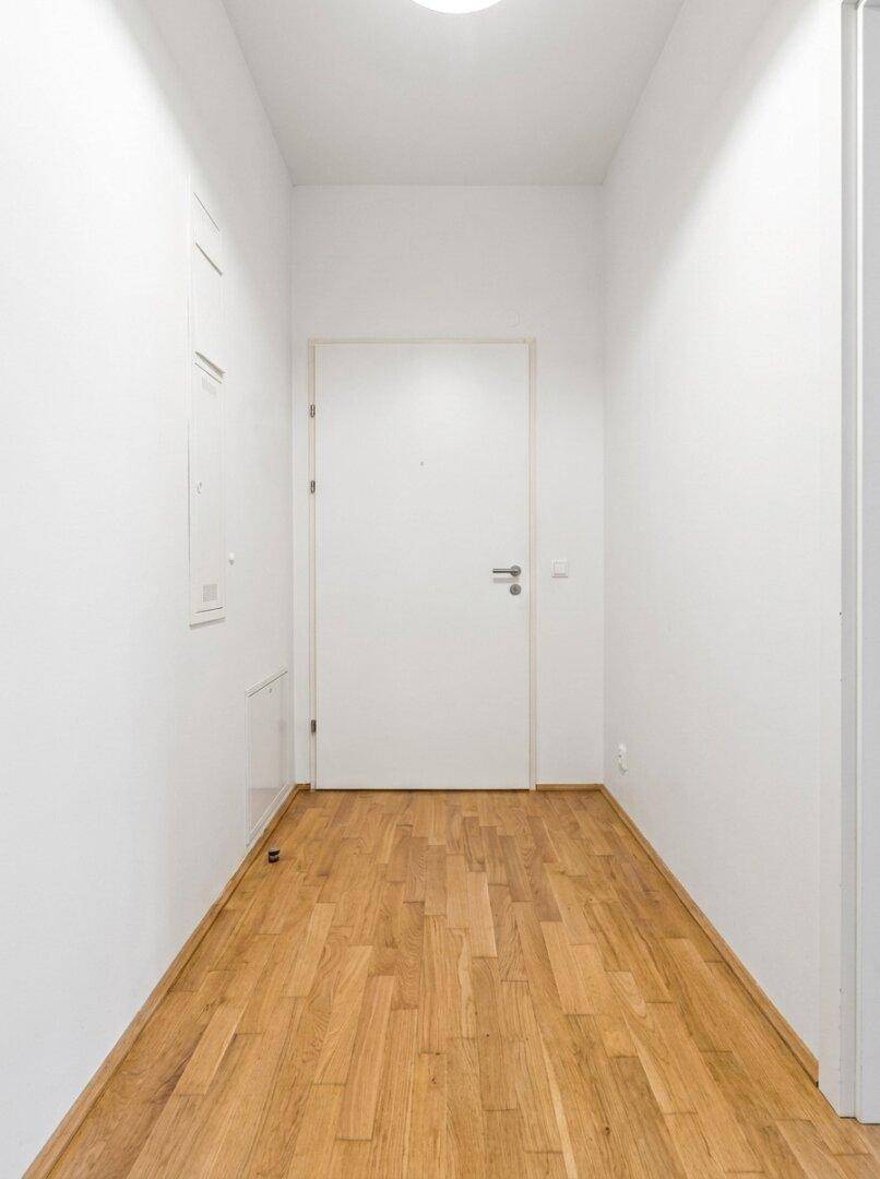 Heller Flur mit weißen Wänden und warmem Holzparkett, führt zu einer weißen Zimmertür.