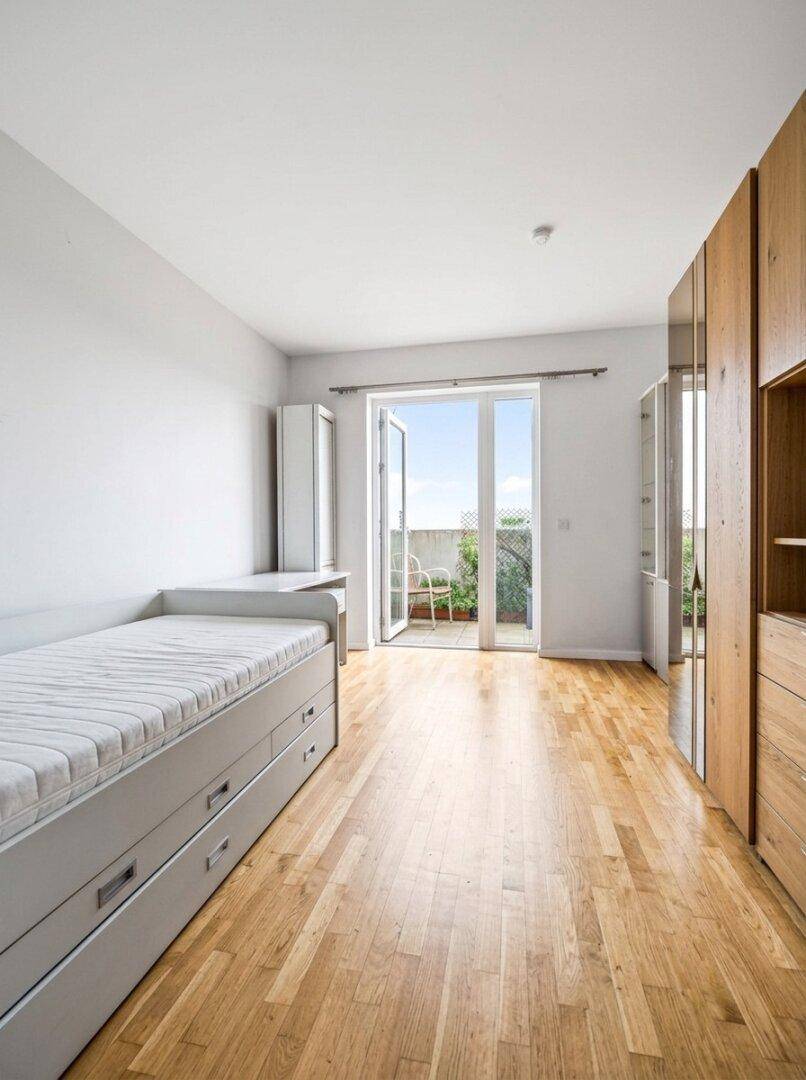 Helles Schlafzimmer mit Einzelbett, Holzschränken und Zugang zu einem sonnigen Balkon.