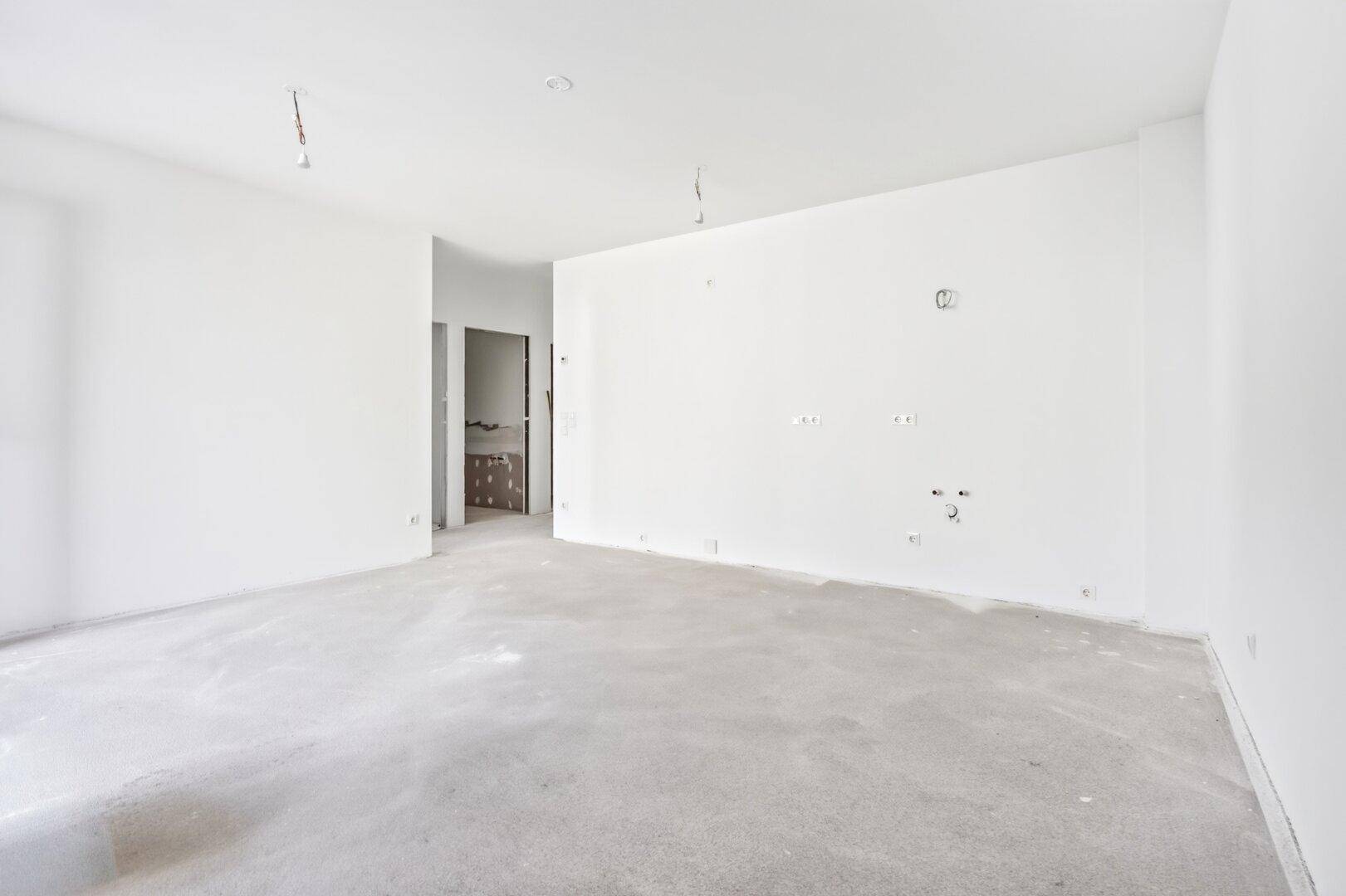 Leerer Raum mit weißen Wänden und Betonboden, vorbereitet für Installationen, mit Durchgang zu weiterem Bereich.