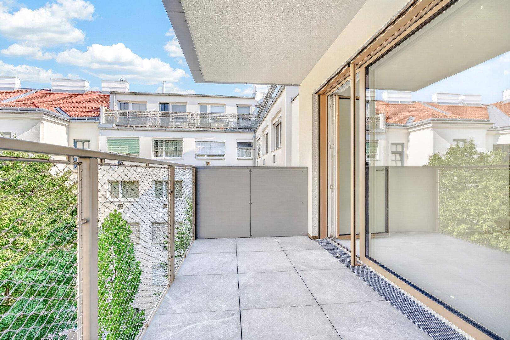 Heller Balkon mit Fliesenboden und großzügiger Glasfront zum Innenbereich, umgeben von Gebäuden.