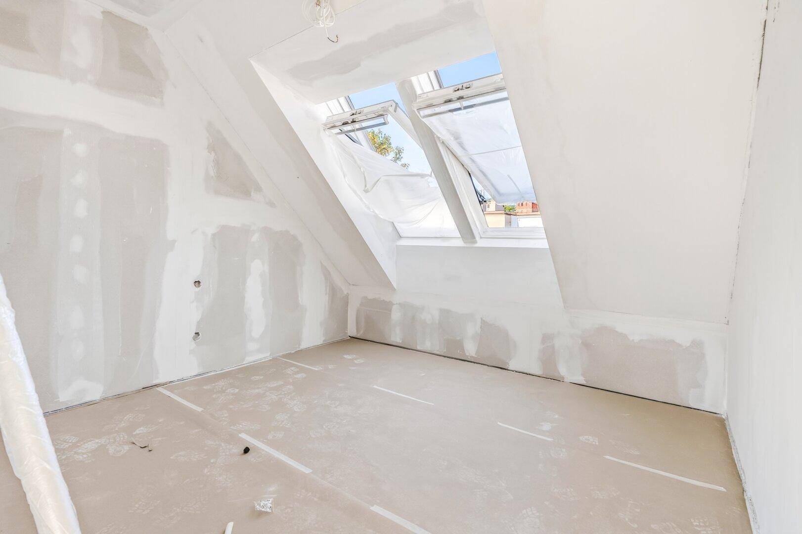 Dachgeschossraum im Rohbau mit schrägen Wänden und zwei großen Dachfenstern für viel Tageslicht.