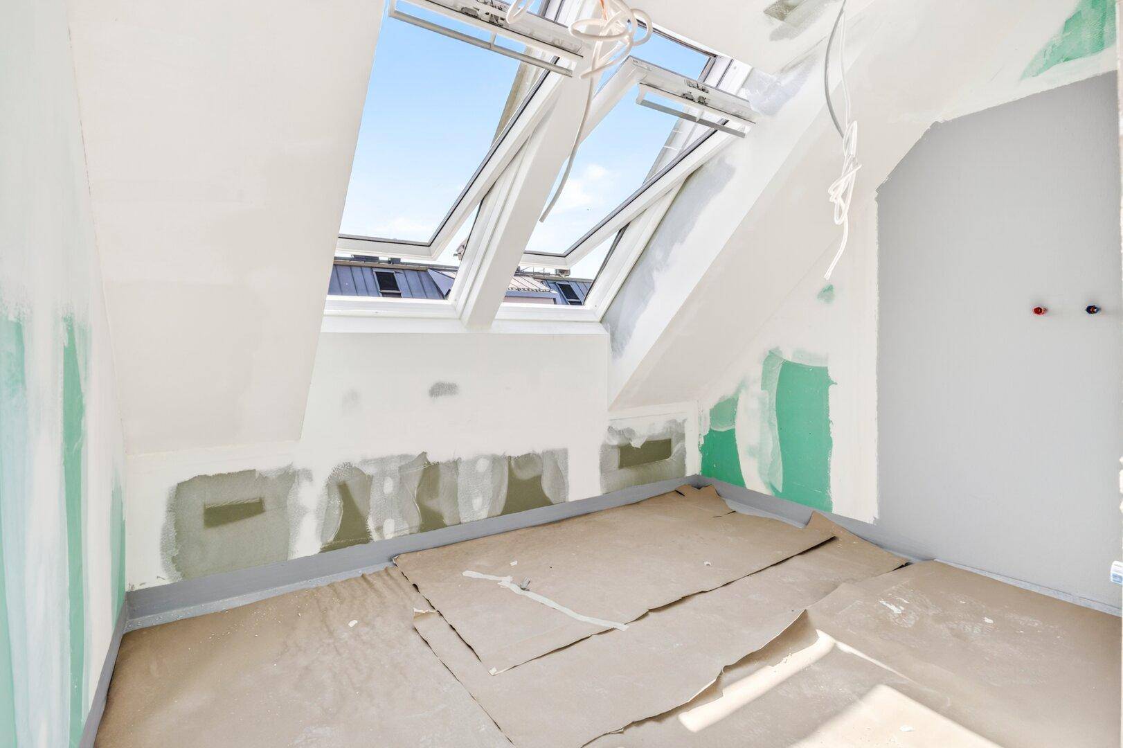 Dachgeschossraum im Rohbau mit schrägen Wänden, Dachfenstern und grünen Trockenbauplatten.
