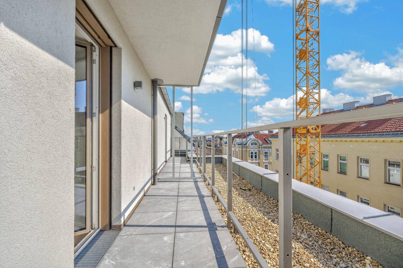 Langer Balkon mit Fliesenboden und Kiesbett, bietet einen seitlichen Blick auf die Stadt und eine Baustelle.