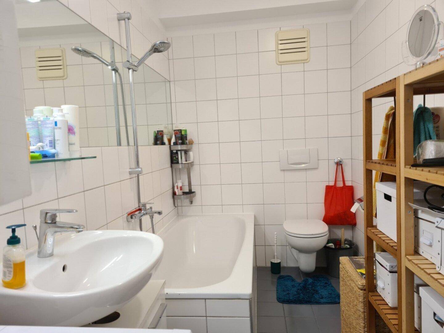 Funktionales Badezimmer mit Badewanne, Duscharmatur, Waschbecken und Holzregal.