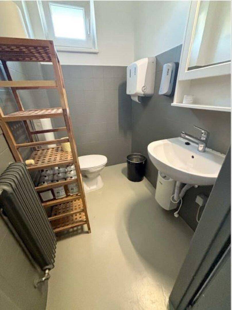 Badezimmer mit grauen Fliesen, Toilette, Waschbecken, Spiegelschrank und einem Fenster für Tageslicht.