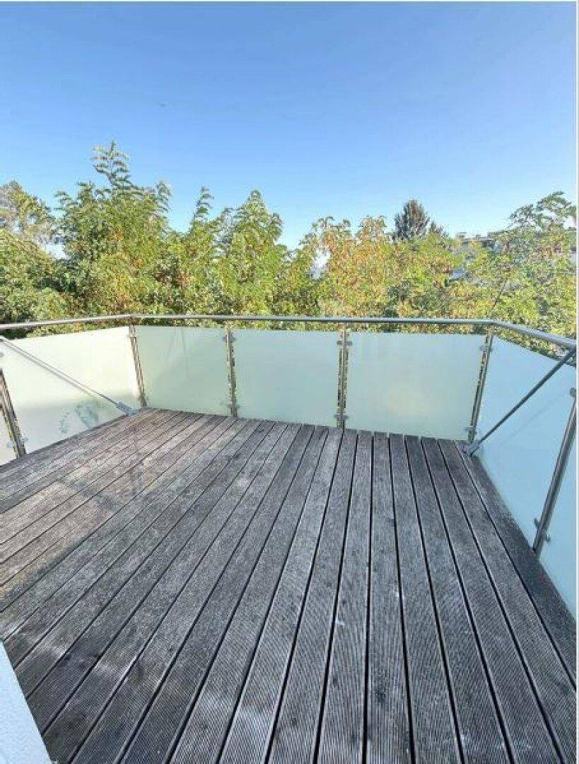 Balkon mit Holzdielenboden und Glasgeländer, umgeben von üppigem Grün und Bäumen.