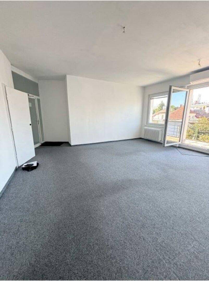 Heller, leerer Raum mit grauem Teppichboden, weißen Wänden und einem offenen Zugang zu weiteren Räumen.