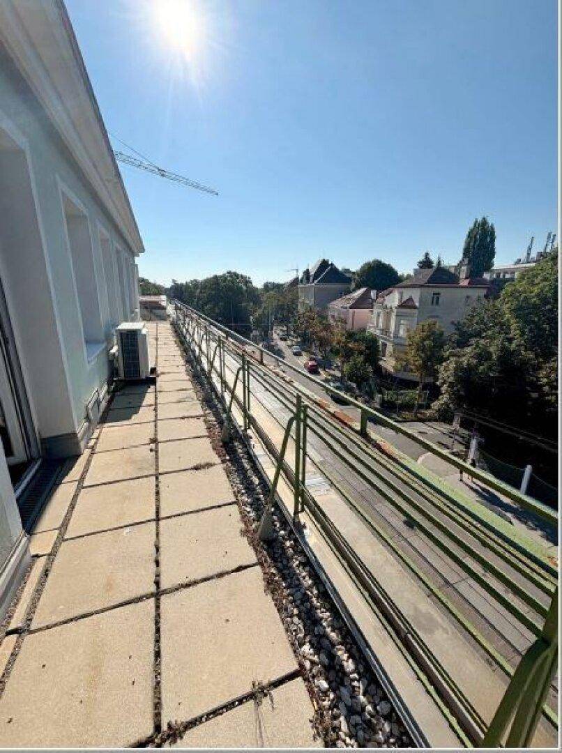 Lange, schmale Terrasse mit Betonplatten und Metallgeländer, bietet Blick auf die Straße und umliegende Gebäude.
