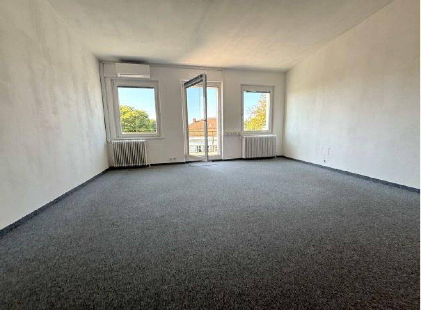 Leerer Raum mit grauem Teppichboden, Klimaanlage und Fenstern, die viel Tageslicht hereinlassen.