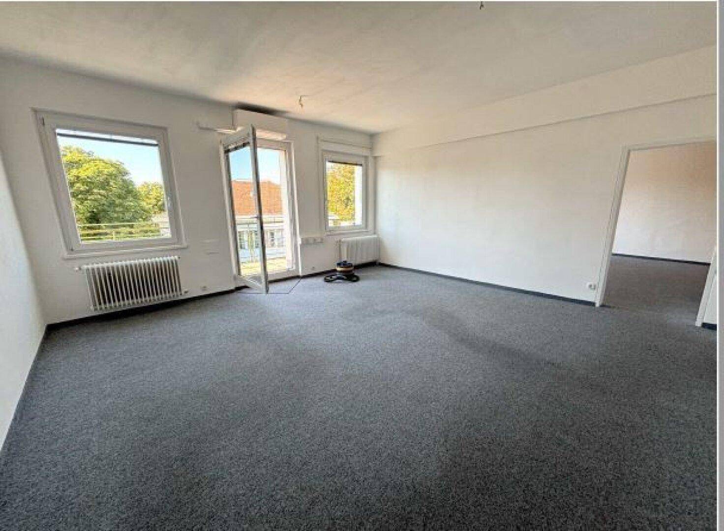 Geräumiger, leerer Raum mit grauem Teppichboden, weißen Wänden und Zugang zu einem Balkon.