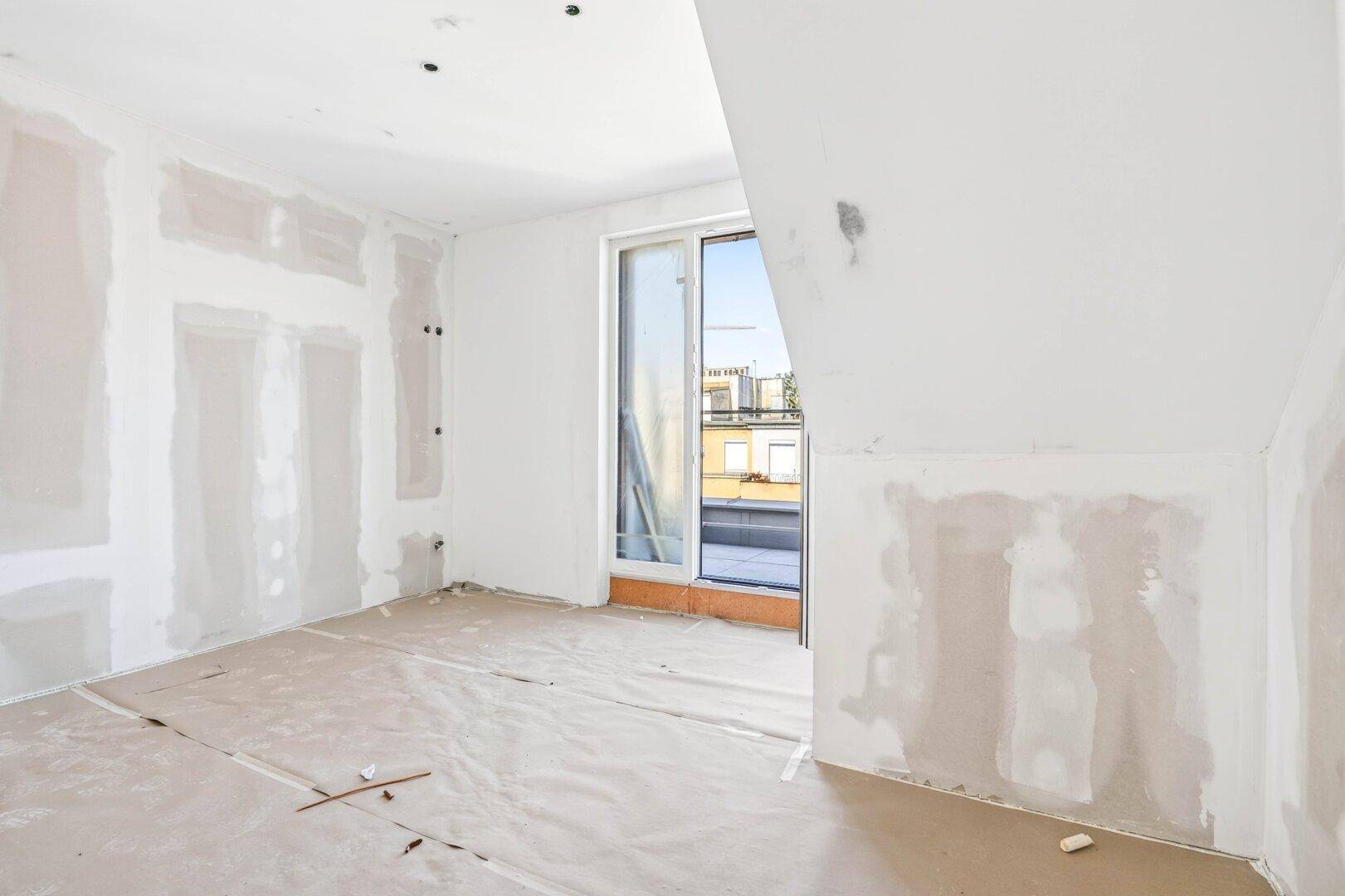 Rohbauzimmer mit Schiebetür zu einem Außenbereich und schräger Decke, viel Tageslicht.