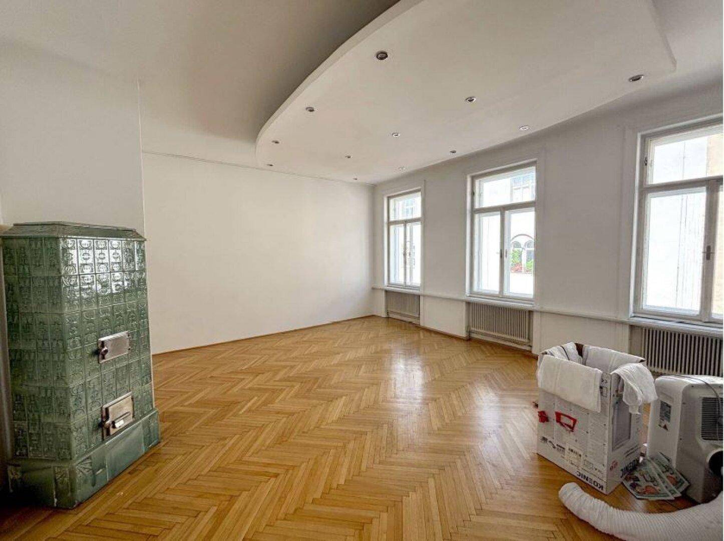 Großer, heller Raum mit Fischgrätparkett, hohen Fenstern und einem traditionellen Kachelofen.