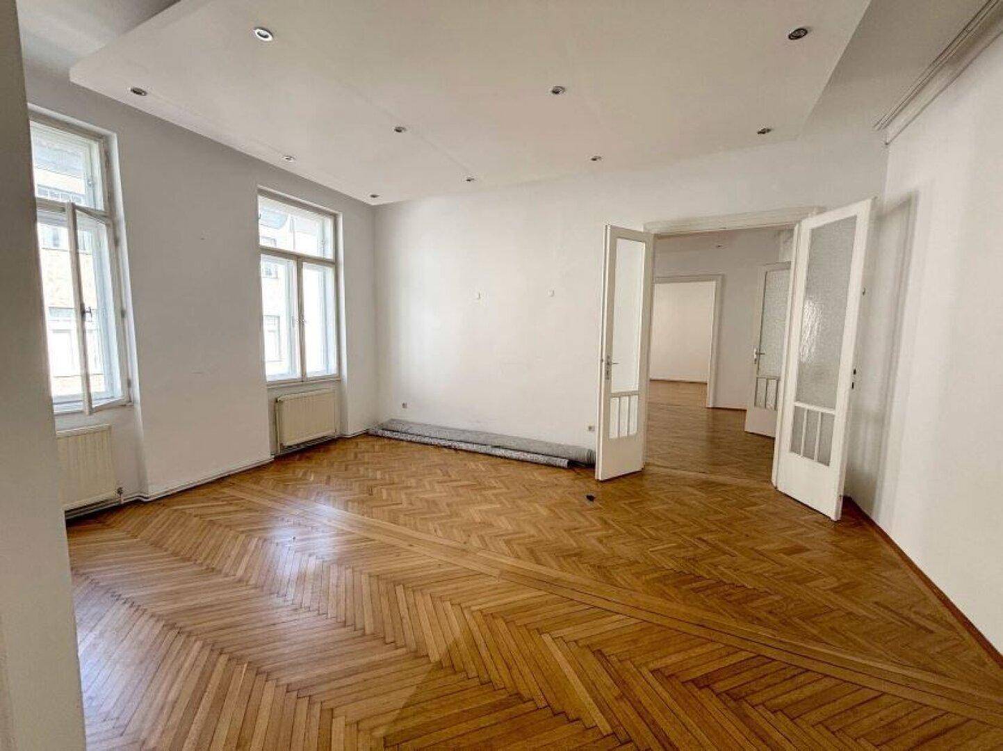 Heller Wohnraum mit Fischgrätparkett, großen Fenstern und einer abgehängten Decke mit Einbaustrahlern.