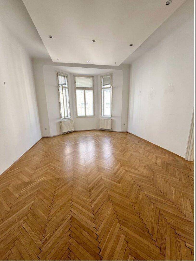 Geräumiger Raum mit Erkerfenstern, Fischgrätparkett und einer modernen Deckenkonstruktion mit Einbaustrahlern.