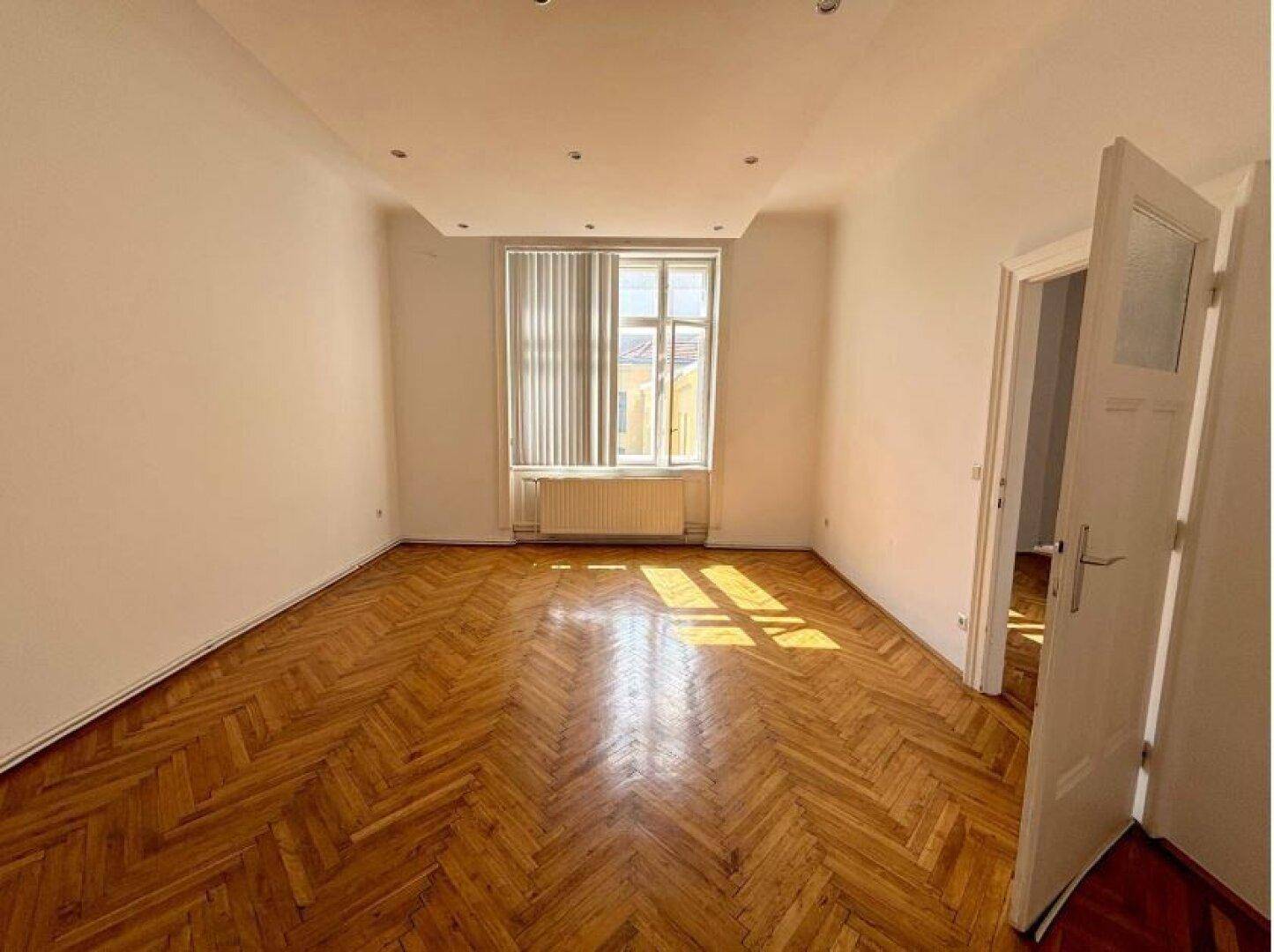 Heller Raum mit Fischgrätparkett, einem großen Fenster mit Jalousien und einer modernen Deckenbeleuchtung.