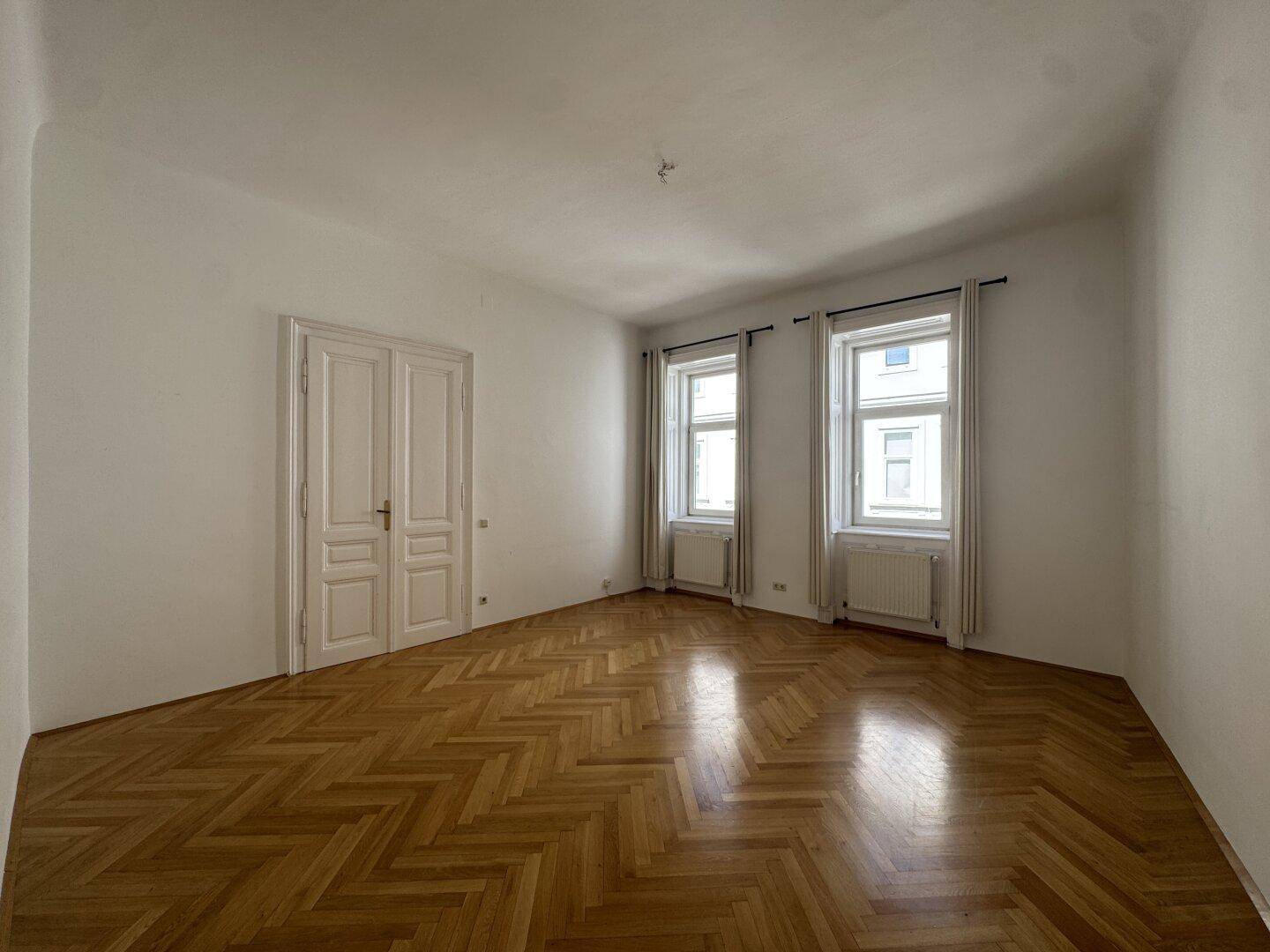 Geräumiges Zimmer mit hellem Fischgrätparkett, weißen Wänden und zwei Fenstern, ideal als Wohn- oder Schlafzimmer.