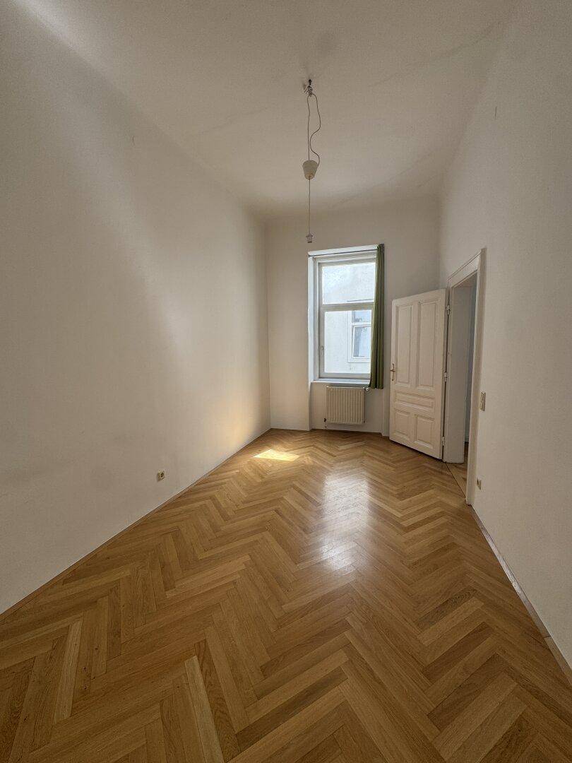 Helles, leeres Zimmer mit einem Fenster, weißen Wänden und einem schönen Fischgrätparkettboden.