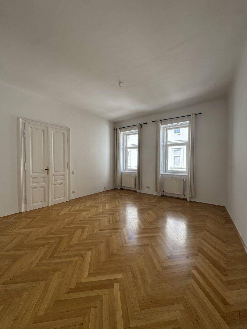 Geräumiges, leeres Zimmer mit weiß gestrichenen Wänden, zwei Fenstern und klassischem Fischgrätparkettboden.