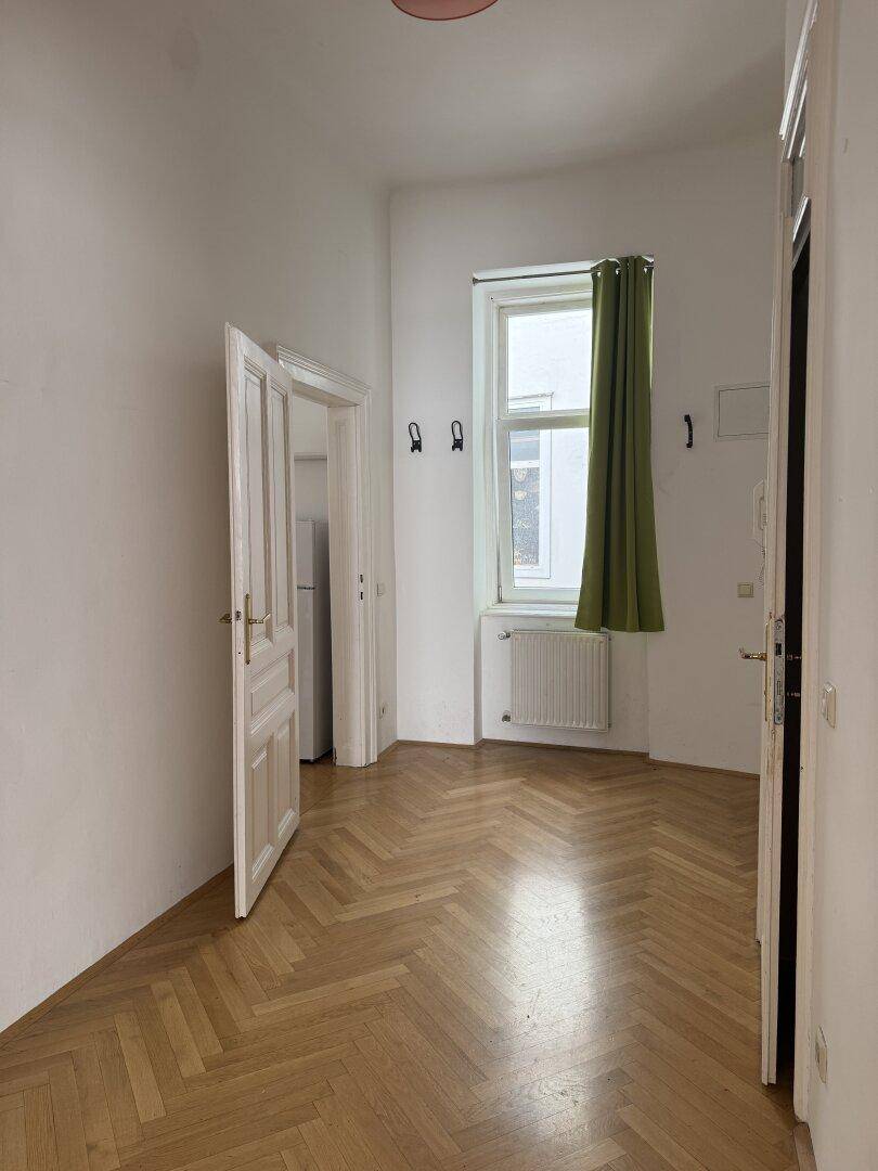 Heller Flur mit Fischgrätparkett und einem Fenster, führt zu einem weiteren Raum mit Kühlschrank.