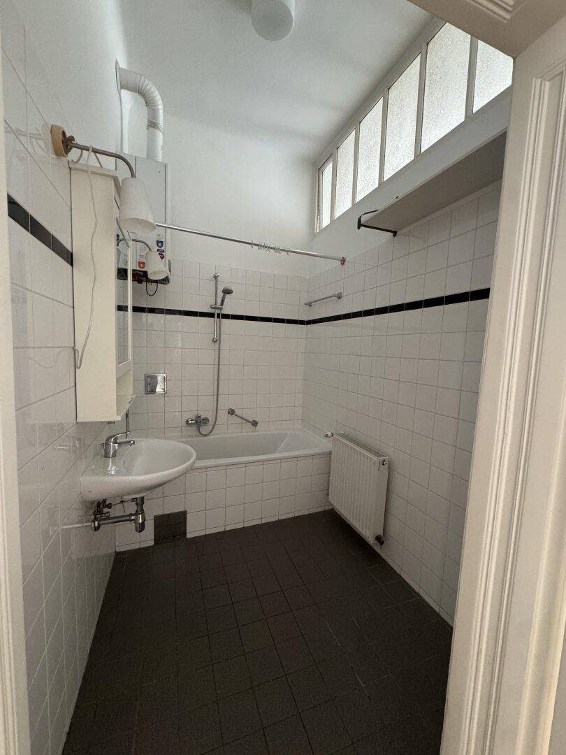 Badezimmer mit Badewanne, Duschfunktion, Waschbecken und weißen Fliesen, verfügt über hohe Fenster für Tageslicht.