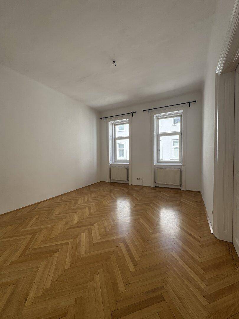 Geräumiges Zimmer mit Fischgrätparkett, weißen Wänden und zwei Fenstern, ideal für vielfältige Nutzung.