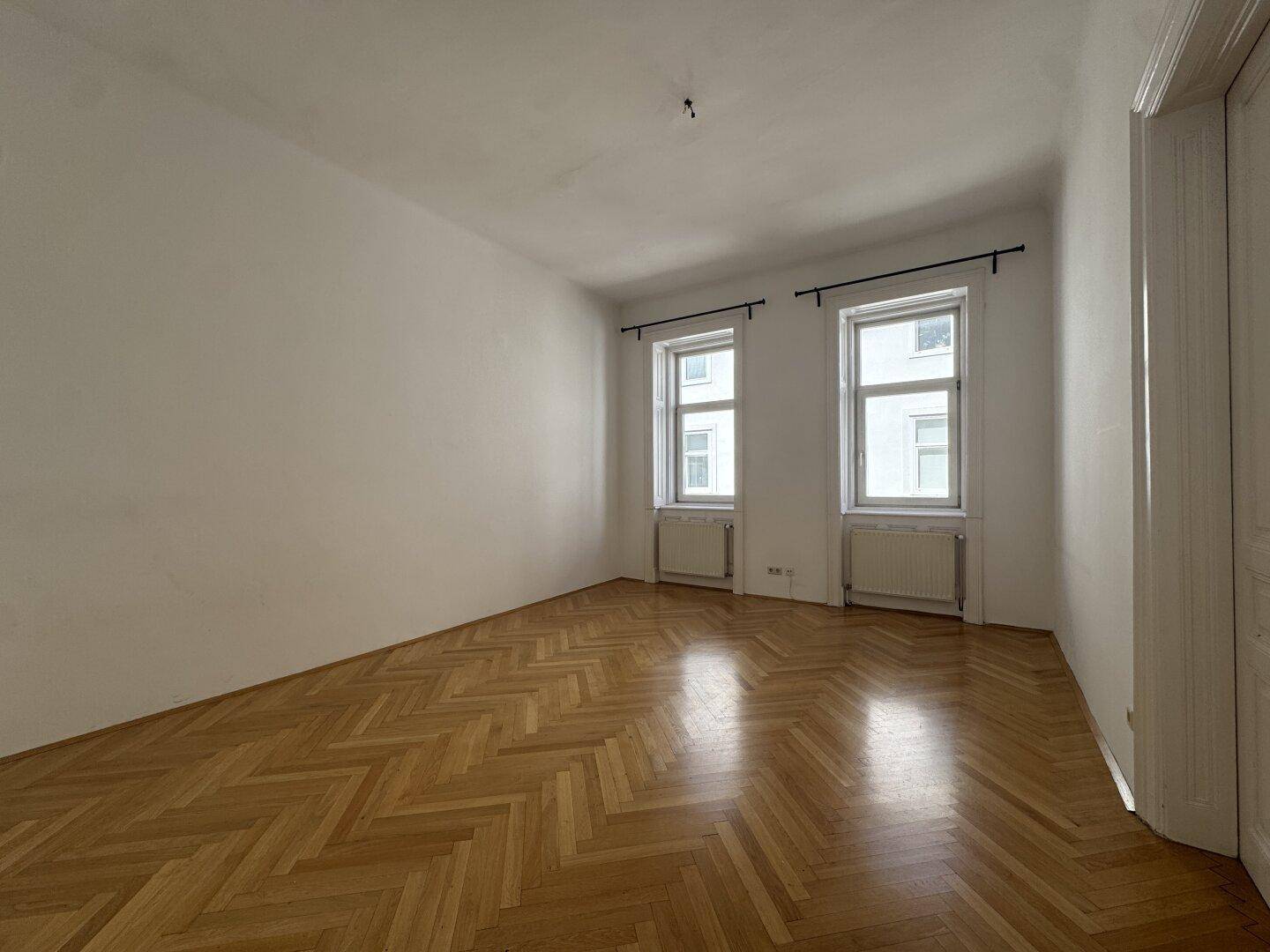 Helles, leeres Zimmer mit zwei Fenstern, weißen Wänden und glänzendem Fischgrätparkettboden.
