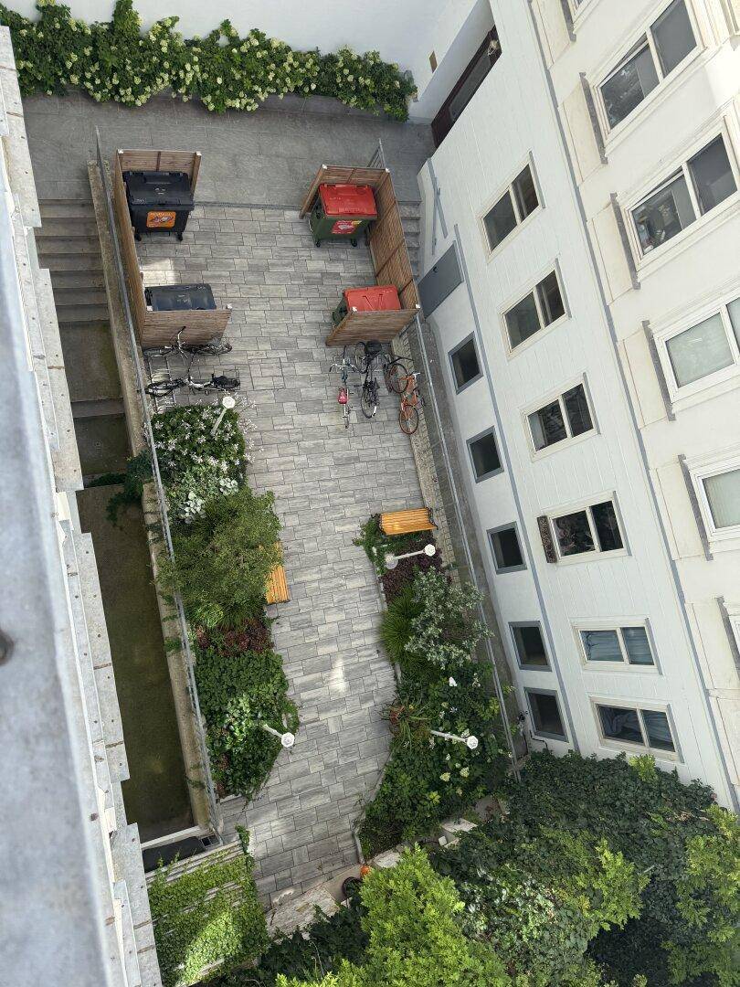 Blick von oben auf einen begrünten Innenhof mit gepflastertem Weg, Bänken und Mülltonnen.