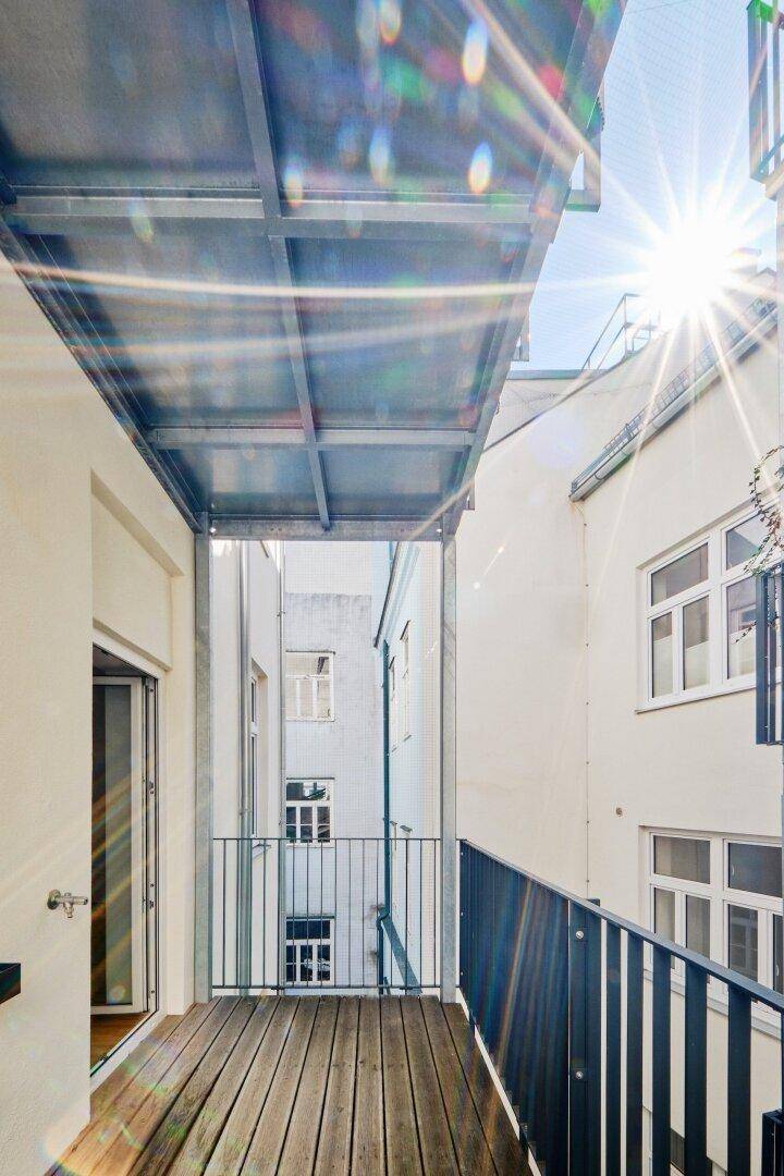 Sonniger Balkon mit Holzbelag und Metallgeländer, der einen Blick in den ruhigen Innenhof bietet.