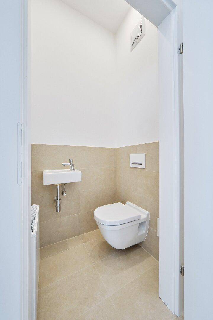 Modernes Gäste-WC mit wandhängender Toilette, kleinem Waschbecken und beigefarbenen Fliesen.