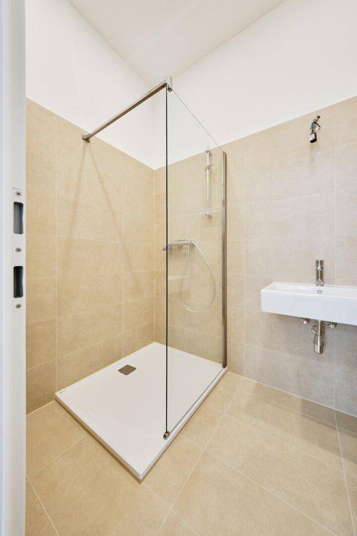 Modernes Badezimmer mit ebenerdiger Dusche, Glasabtrennung und kleinem Waschbecken, alles in hellen Beigetönen.