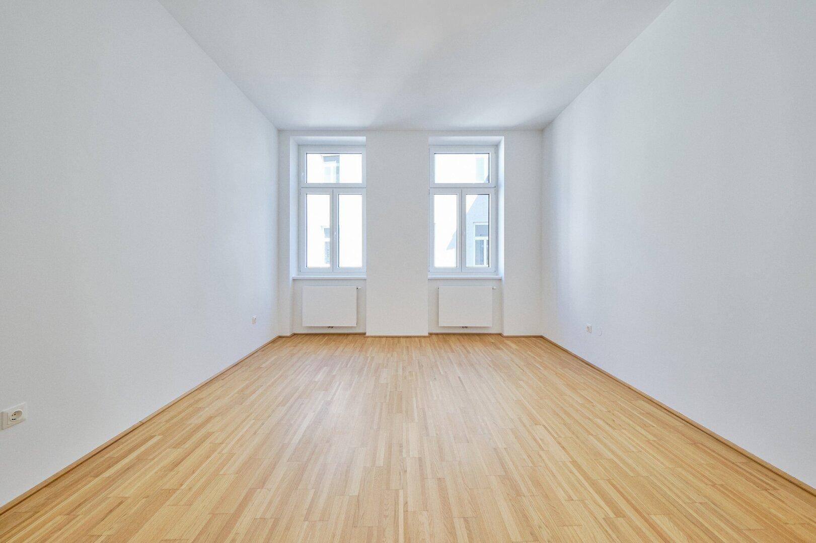 Geräumiges, leeres Zimmer mit zwei großen Fenstern, Parkettboden und weißen Wänden, sehr hell.