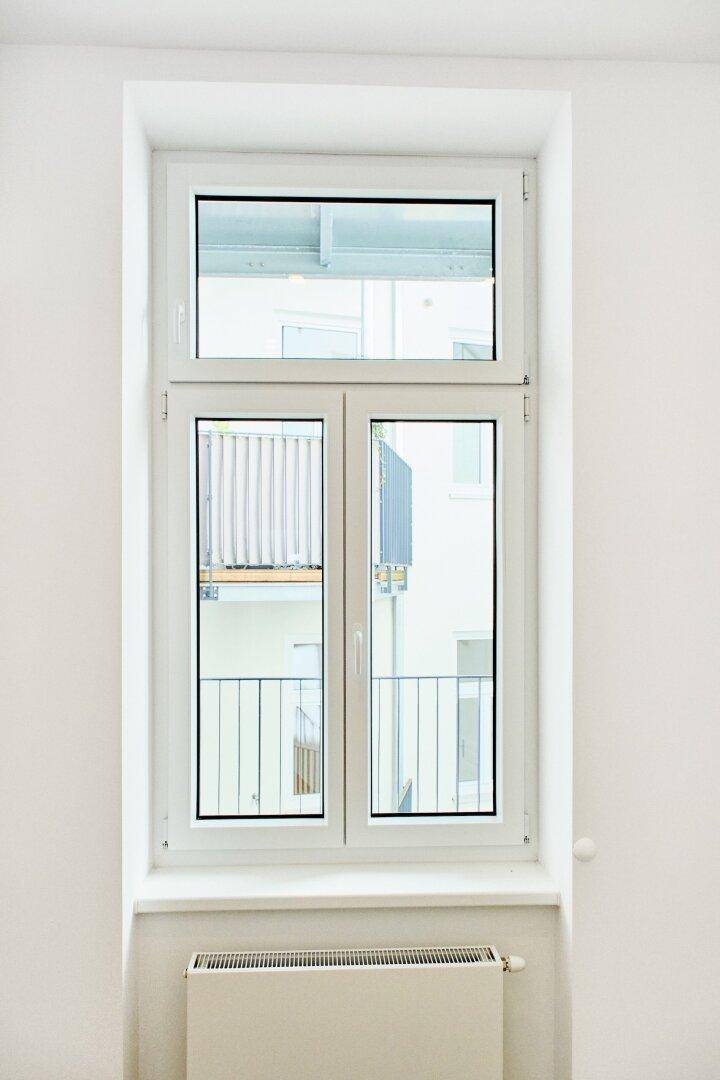 Großes Fenster mit weißem Rahmen, das einen Blick auf die umliegenden Gebäude und Balkone freigibt.